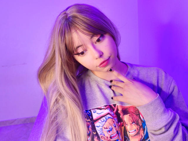 Arieyaara with long blonde hair, wearing a gray sweater with anime print, purple lighting, black nail polish, and a slightly confused expression, touching her chest.