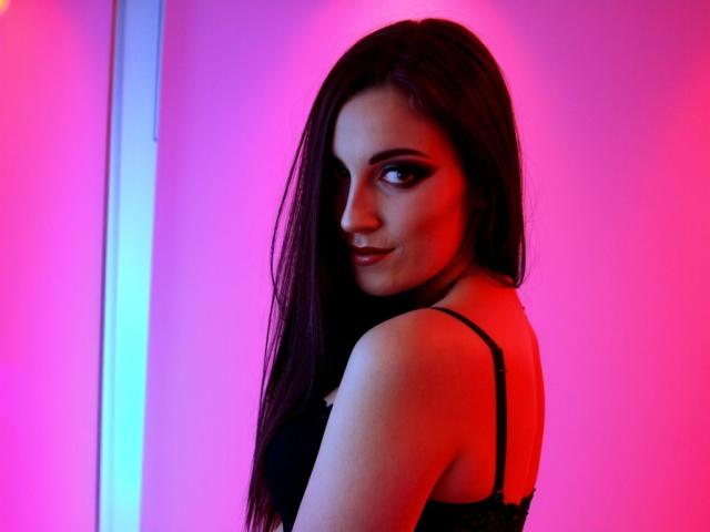 AmandaChilli with long dark hair, looking over her shoulder, smiling, wearing a black spaghetti strap top, illuminated by pink and blue lights. Her tits are slightly visible.