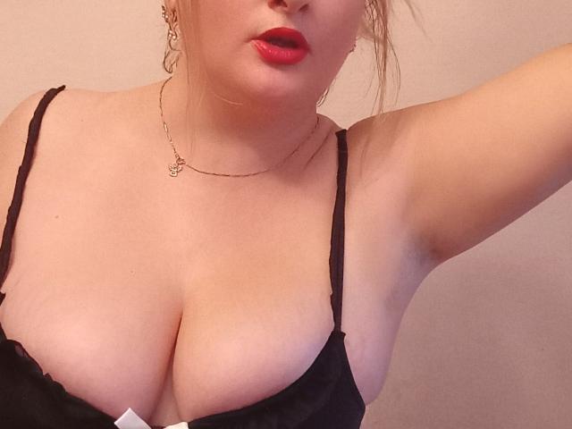 KelliRedhaer with red lips, black bra, and gold necklace, showing her large tits and fair skin, arm raised, blonde hair in messy bun, subtle makeup, soft lighting.