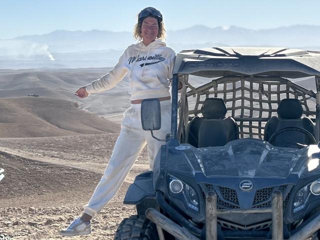 BlackAngeel stands in front of a blue quad bike, wearing a white hoodie and pants, with white sneakers, and a black cap, in a dusty, hilly landscape.