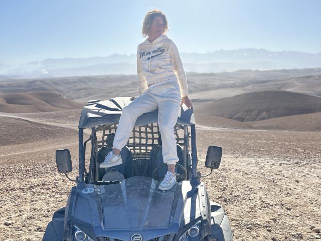 BlackAngeel sitting on a blue ATV with a metal grid cover, wearing a white hoodie and pants, white sneakers, in a dusty, sunlit desert.