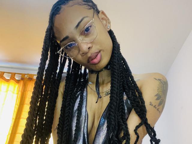 ElaHott, con trenzas negras largas, usando gafas, un vestido negro brillante y un collar, tiene tatuajes pequeños y está sacando la lengua. Sus senos son visibles..
