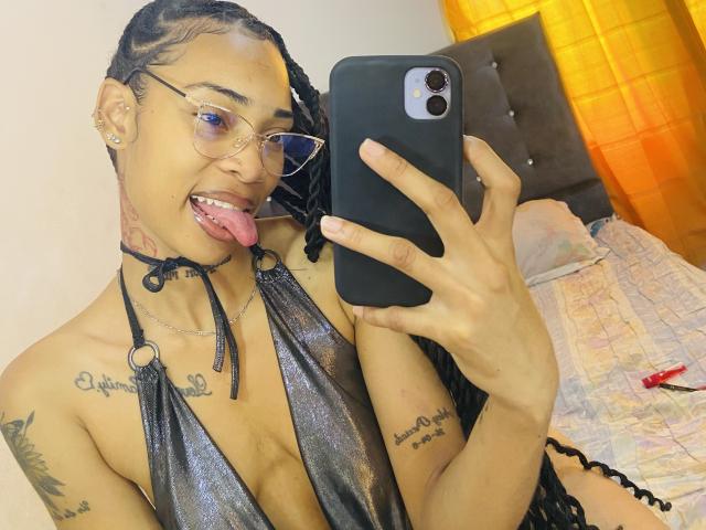 ElaHott with glasses, tongue out, wearing a shiny black halter top, holding a phone, showing tattoos, and braided hair, taking a selfie, with visible cleavage.