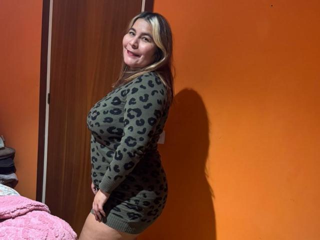 LouiseSexBigBoobs with blonde highlights, smiling, wearing a green leopard-print dress, standing in an orange-walled room, showing her ass, standing side-on, looking over her shoulder.