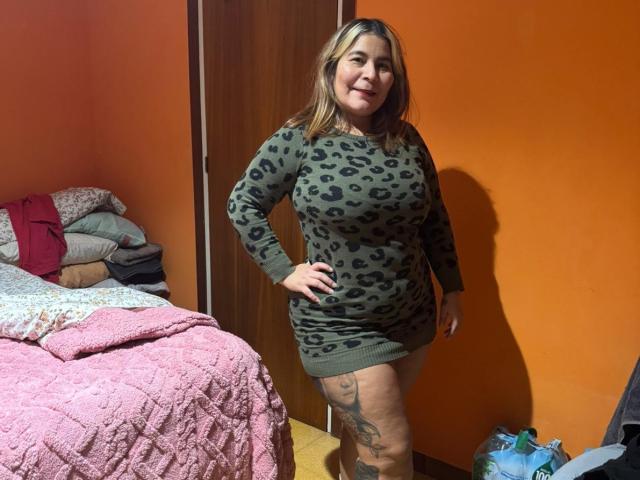 LouiseSexBigBoobs standing in a room, wearing a green leopard-print dress, showing her ass and a tattoo on her right thigh, smiling with hand on hip.