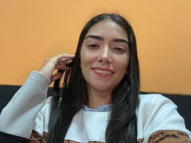 SharonKlart with long black hair, smiling, wearing a white textured shirt, hand near ear, orange background, cute face, slightly shiny skin, relaxed expression.
