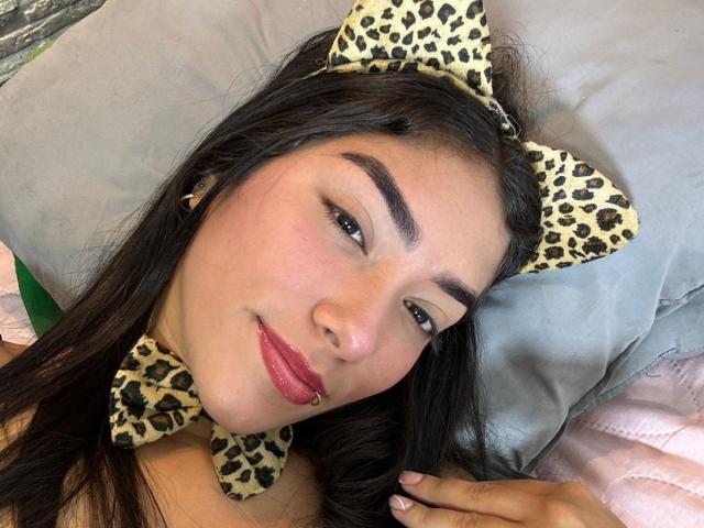 SharonKlart has dark brown hair, a nose ring, and is wearing leopard print cat ears and bowtie, lying on a gray pillow with pink bedspread.
