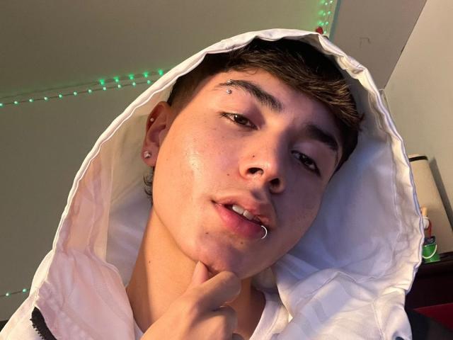 ImJuanda with a white hoodie, piercing in lip, nose, and eyebrow, short brown hair, and ear studs, touching chin, with green LED lights in background.