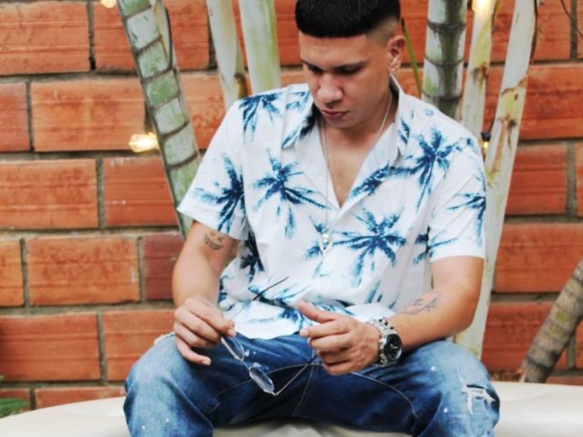 Brian Cruz, avec ses cheveux noirs courts, portant une chemise bleue à motifs de palmiers et un jean bleu, est assis sur une surface blanche, regardant son téléphone, et on peut voir des tatouages sur son bras..