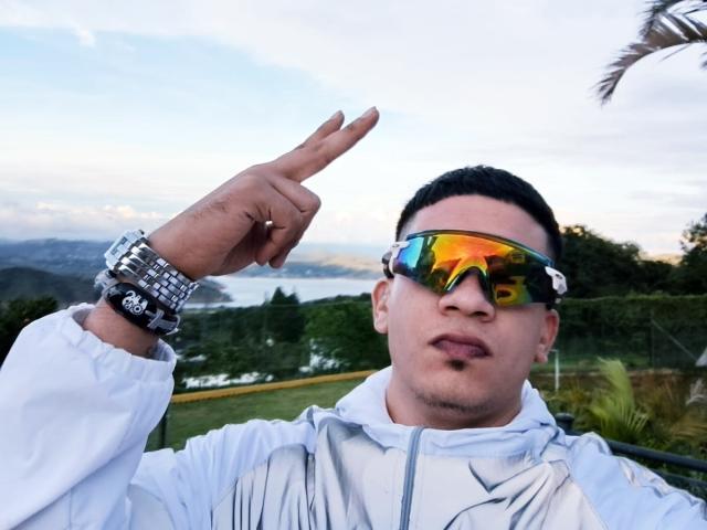Brian Cruz, avec un sourire narquois, portant des lunettes de ski réfléchissantes et une veste blanche, pointe vers le haut avec son index, et porte une montre argentée à son poignet droit..