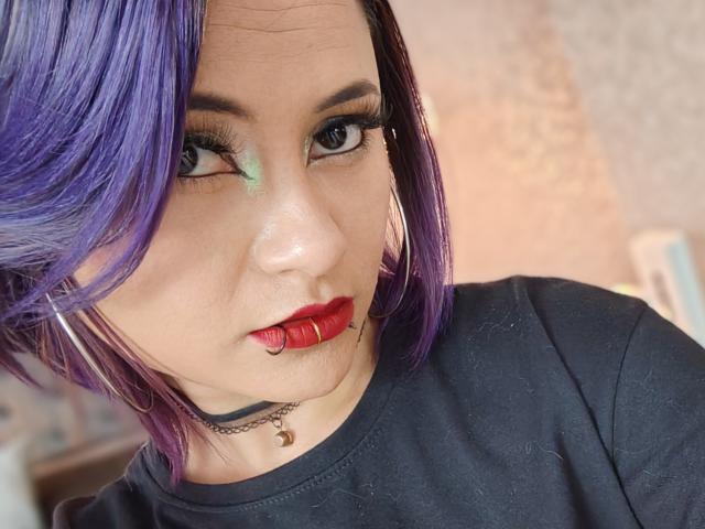 CassieJones with purple hair, green eyeshadow, red lipstick, and a black shirt. She's wearing a choker and has hoop earrings, looking at the camera with a serious expression.