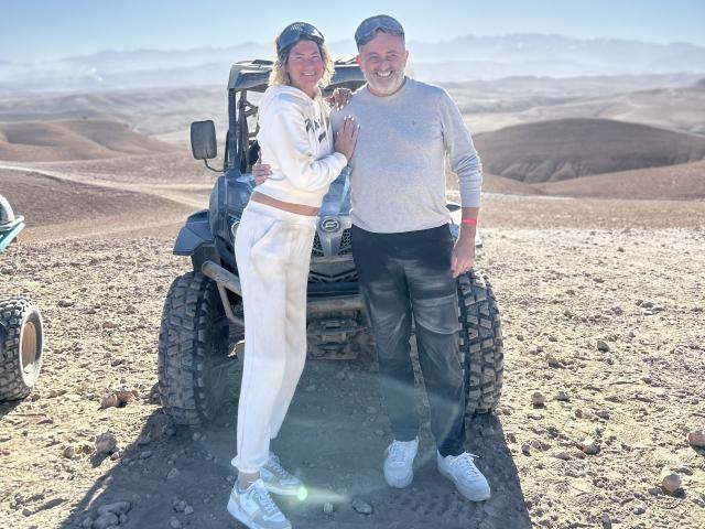 AngeelCouple with tits covered in a white hoodie and white pants stands next to a bearded dude in a gray sweater and black pants, both smiling by a jeep.