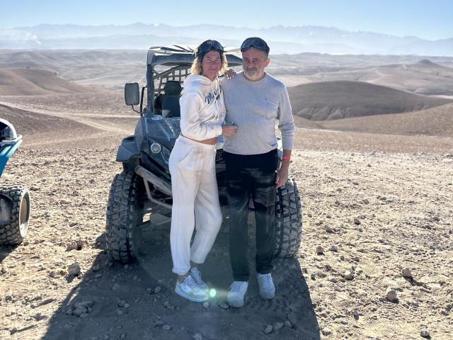 AngeelCouple standing in a desert with a jeep, AngeelCouple in white outfit and sneakers, AngeelCouple in gray sweater and black pants, both smiling, AngeelCouple's hands on jeep.