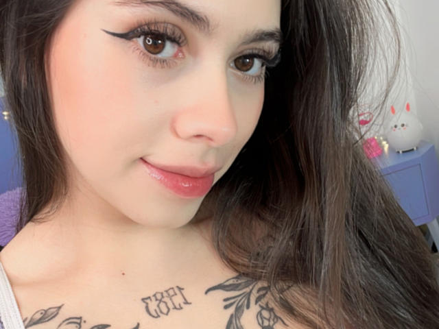 ChanelDeRosee tem pele clara, olhos castanhos, cabelos longos e escuros, delineador preto, lábios rosados e brilhantes, e tatuagens no peito. Ela está sorrindo levemente, mostrando um rosto bonito e sexy..