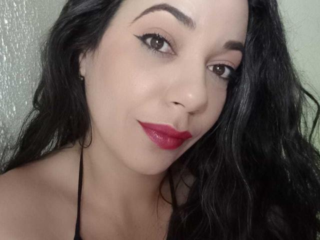 YolittS has dark, wavy hair, brown eyes, and red lipstick. She's smiling with black eyeliner, wearing a black halter top, and has small earrings.