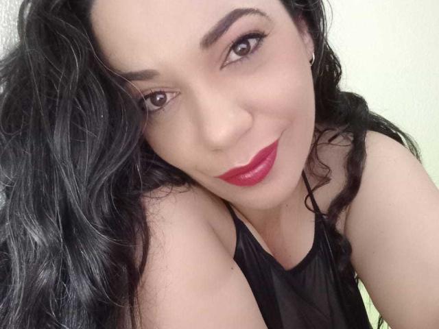 YolittS with dark wavy hair, red lipstick, and black sleeveless top, smiling at the camera, showing her shoulder and upper chest. Boobs slightly visible. Soft, cute face.