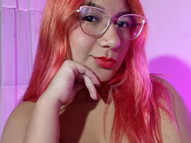 AradiaX with red hair, glasses, and red lipstick, resting her chin on her hand, showing her tits. She's wearing a nose ring and has a cute, confident look.