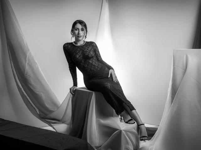 Black-and-white photo of AlanaBazan, sitting on a draped table, wearing a lace long-sleeve bodysuit, showing her tits, with her legs crossed, and wearing strappy heels.