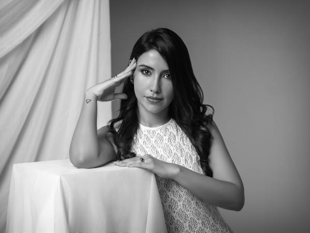 AlanaBazan with long dark hair, lace dress, and a ring, leaning on a white-covered table, hand on head, looking at camera, grayscale photo.
