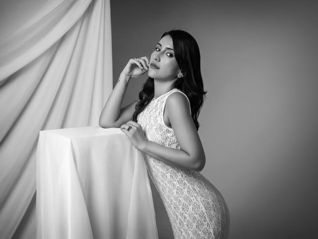 AlanaBazan in a lace dress, leaning on a white table, looking at the camera, with one hand near her mouth, her dark wavy hair flowing down. Black and white.