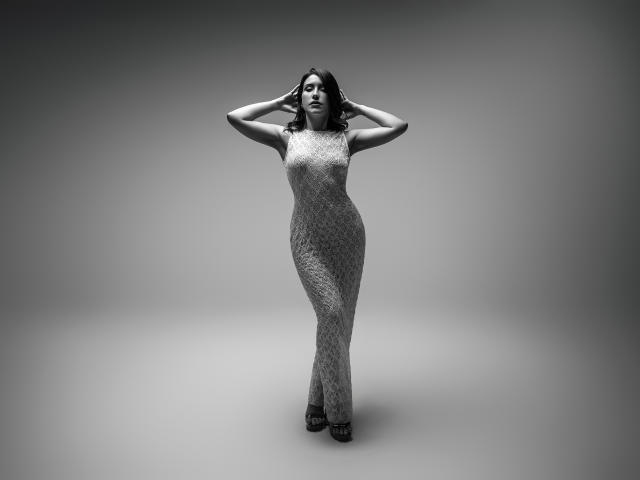 AlanaBazan stands in a grey, patterned, sleeveless dress, hands behind her head, with a confident, sultry expression. She has dark, wavy hair and is wearing black heels.