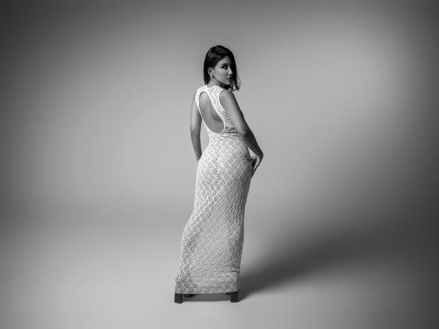 AlanaBazan stands in a black-and-white photo, wearing a sleeveless, lace, floor-length dress, looking over her shoulder with her hands on her pregnant belly, showing her ass.
