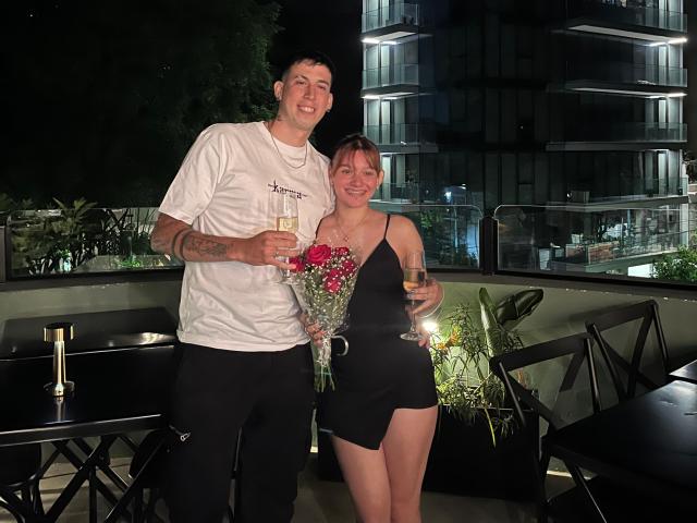 LuluCouple, ein Mann mit Tattoos und einem weißen T-Shirt, steht neben LuluCouple, einer Frau in einem schwarzen Kleid, die Blumen und Champagner hält und lächelnd in die Nacht blickt..