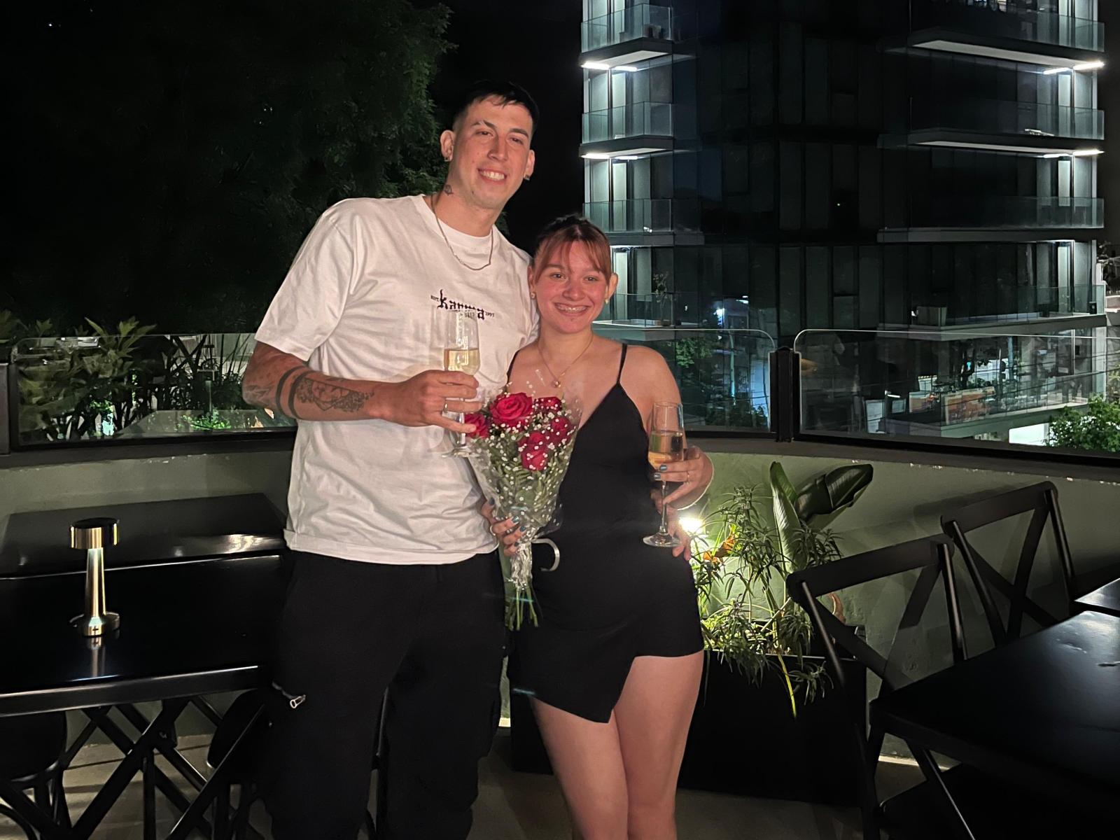 LuluCouple, ein Mann mit Tattoos und einem weißen T-Shirt, steht neben LuluCouple, einer Frau in einem schwarzen Kleid, die Blumen und Champagner hält und lächelnd in die Nacht blickt..