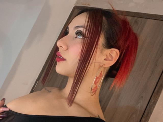 ValeryHarllow has red-tipped black hair in a ponytail, large hoop earrings, and pink lipstick, with blue eyes and a tattoo on her neck, wearing an off-shoulder black top.