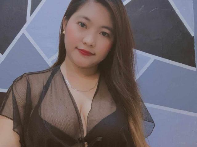 BettyStar with long brown hair, wearing a sheer black top, showing her big tits, smiling, red lipstick, and a silver necklace, against a geometric patterned wall.