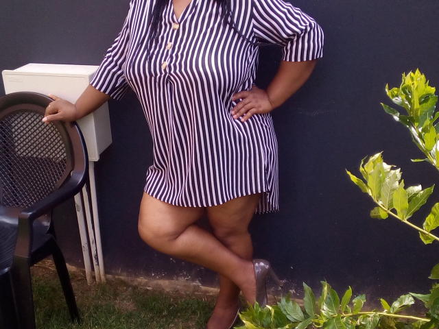 KillerBoobsLona in a striped shirt, her tits visible, standing with one leg crossed, hand on hip, next to a chair and air conditioning unit, green plant in foreground.