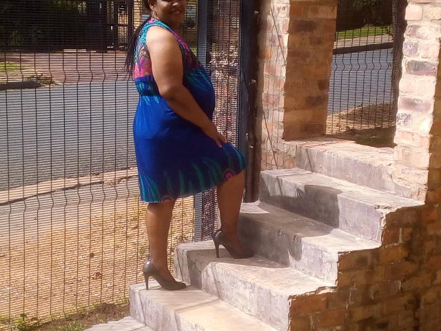 KillerBoobsLona standing on concrete stairs in a blue dress with pink and green patterns, brown high heels, brick wall on the right, and metal fence in the background.
