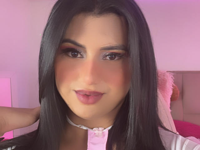 LolaOberle has long black hair, pink eyeshadow, and red lipstick. She's wearing a white collar with a pink button, and her hand is touching her hair.
