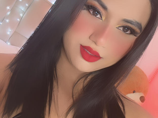 LolaOberle has dark hair, red lipstick, and thick eyelashes. She's looking at the camera with a slight smile. Her makeup is bold, with pink blush on her cheeks.