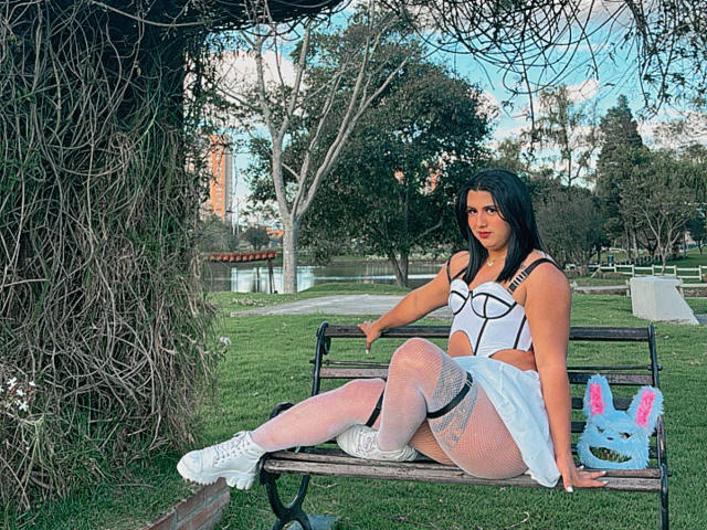 LolaOberle sits on a bench, wearing white fishnet stockings, a white crop top with cutouts, and a white skirt. Her black hair is down, and she's looking at the camera.