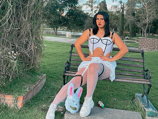 LolaOberle with black hair, red lipstick, white cut-out top, sheer white skirt, white stockings, bunny slippers, sitting on bench, hands on thighs, park, evening light.