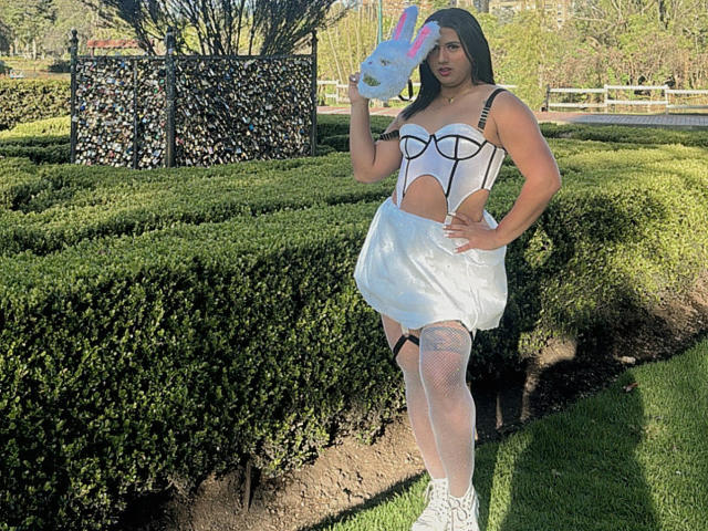 LolaOberle with long black hair, white cut-out dress, black straps, white fishnet stockings, white sneakers, holding pink and white bunny mask, standing in green garden.