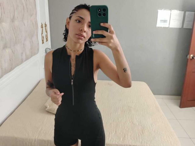SilvanaXOwen in a tight black sleeveless zip-up dress, holding a green phone, short curly hair, small tattoo on right arm, necklace, standing in a bedroom, looking at mirror.