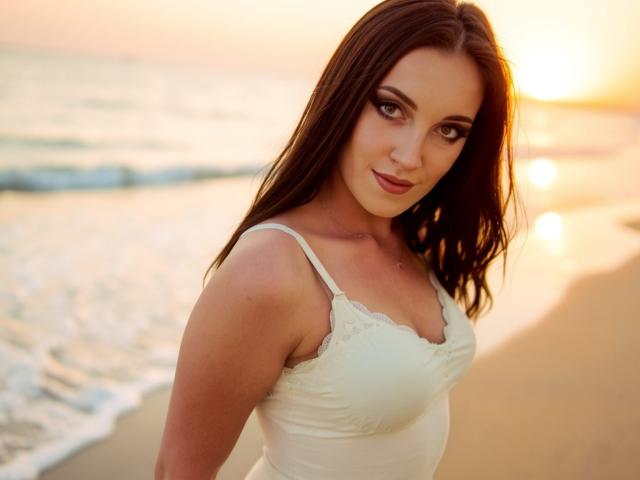 AmandaChilli with brown hair, white lace top, and small tits, standing on a beach at sunset, looking at the camera with a slight smile.