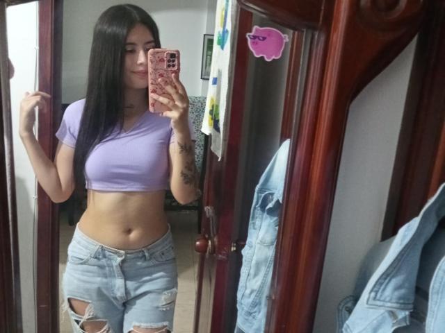 AlisonKweel with long black hair, purple crop top, showing her abs, and ripped blue jeans, holding a pink phone in a mirror selfie, showing her tattoos.