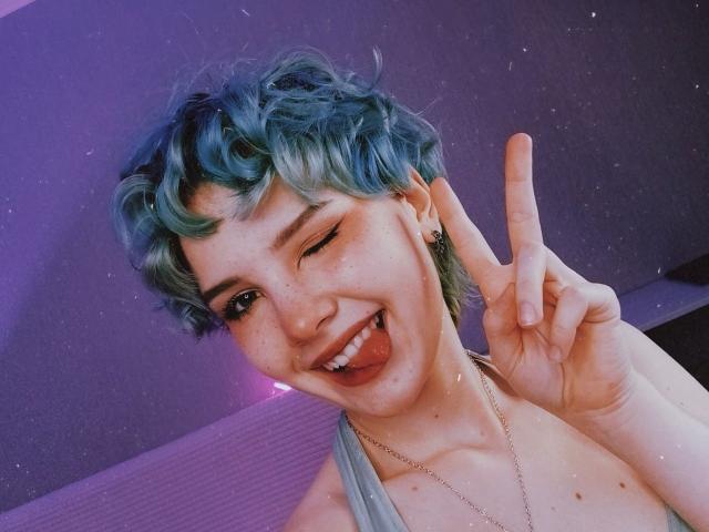 ShellyLove69 with short, curly blue-green hair, winking, sticking out tongue, and flashing a peace sign. She's topless, showing small tits, and has a cute, freckled face.