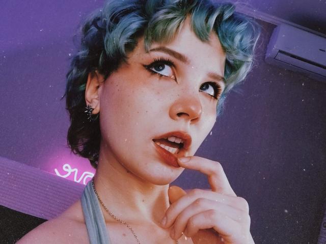 ShellyLove69 with short, curly blue-green hair, freckles, and blue eyes, biting her finger, looking up with half-open lips, wearing a grey top, and a necklace.