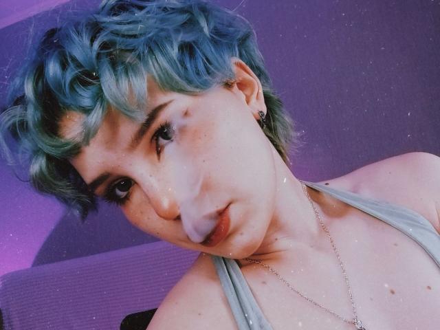 ShellyLove69 has short, curly blue-green hair, freckles, and a silver necklace. She's wearing a grey tank top, with a serious, slightly pouty expression. Her tits are small.