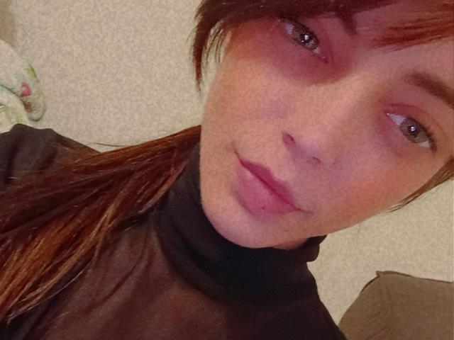 MaitresseWolfly with red hair, blue eyes, and pink lipstick, wearing a black sheer turtleneck, has a subtle smile, and her brown ponytail is visible.
