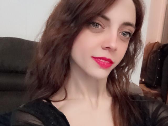 MaitresseWolfly has pale skin, brown wavy hair, and bright pink lipstick. She's wearing a black sheer top, looking at the camera with a slight smile.