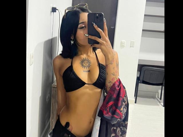 SalomeMille with black bikini, sun tattoo on chest, arm tattoos, holding phone, black hair, slim, tan skin, sexy, mirror selfie, black pants, room with door and chair.