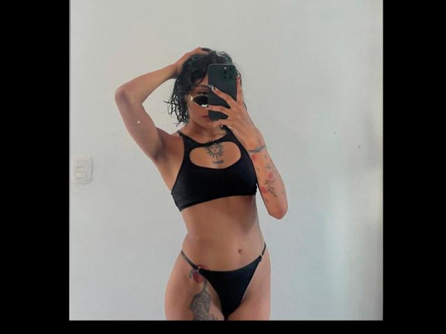 SalomeMille in black sports bra and thong, holding phone, covering face, wavy black hair, tattoos on arm and thigh, hand in hair, slim, small breasts, light skin.