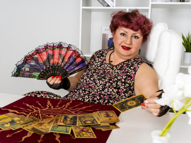 GrannyEva met krullend roze haar, rode lippenstift, een bloemenjurk, een zwarte handschoen zonder vingers, die een waaier vasthoudt, zittend aan een bureau met tarotkaarten en een witte stoel. Rode nagels, een witte bloem..