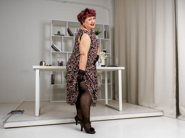 GrannyEva with curly pink hair, floral dress, black lace gloves, sheer black stockings, and black heels, showing her ass and tits, standing in front of a white desk.