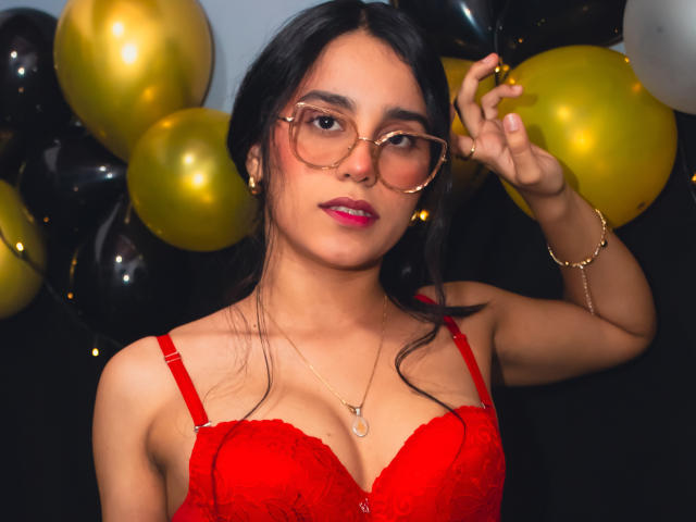 TiffannyRose con gafas, sostén de encaje rojo, lápiz labial rosa, sosteniendo gafas, pulsera dorada, collar, globos negros y dorados, busto medio, ojos marrones, cabello negro largo, sexy, mirada segura..